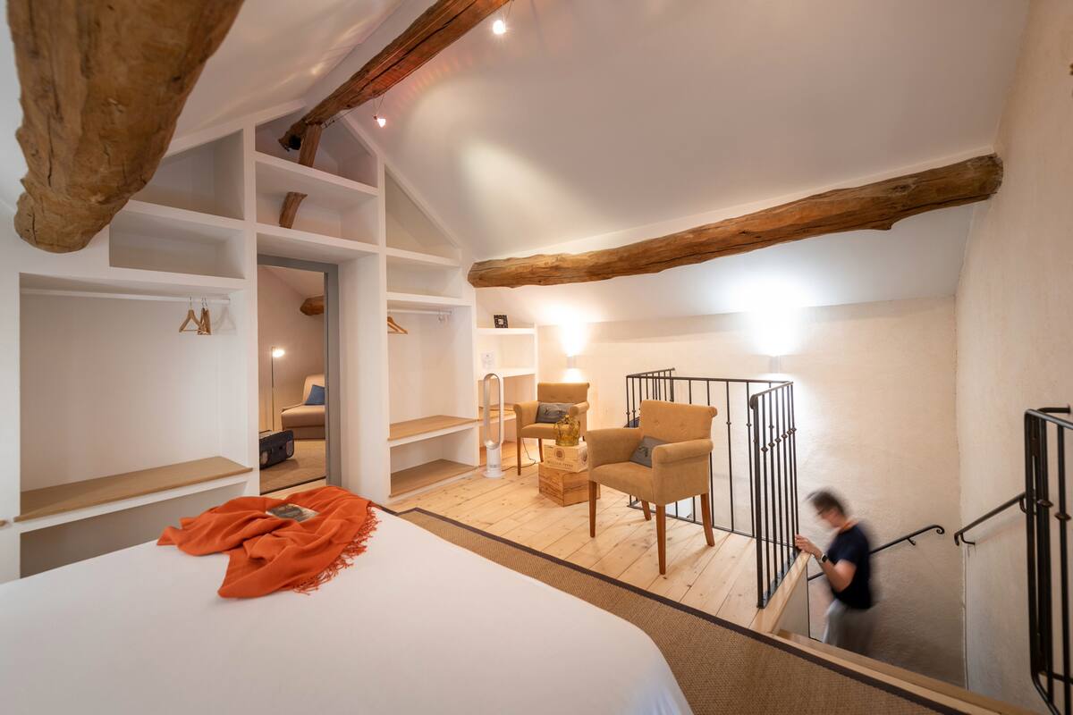 A bedroom with a large bed, wooden beams, built-in shelving, a chair, and a staircase.