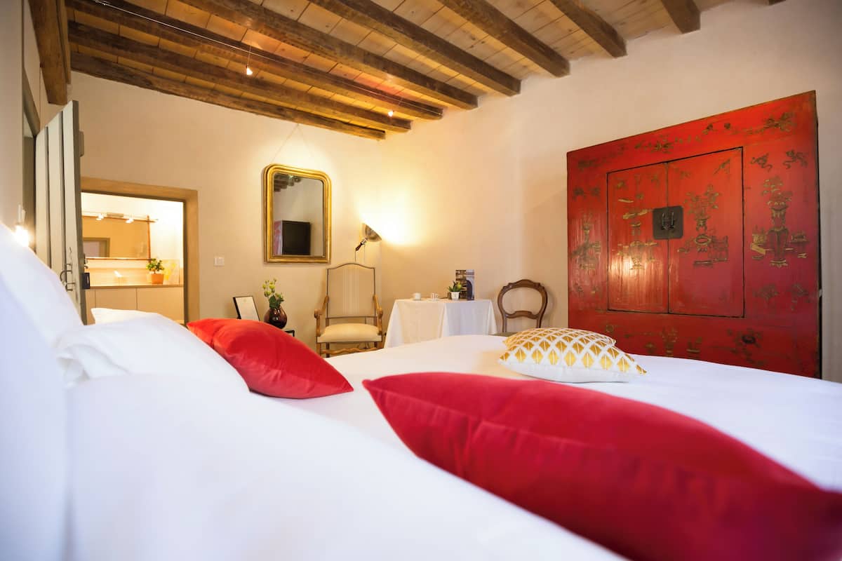 A hotel room with a bed, red pillows, a wooden ceiling, a mirror, a table with a lamp, and a red door with decorative panels.