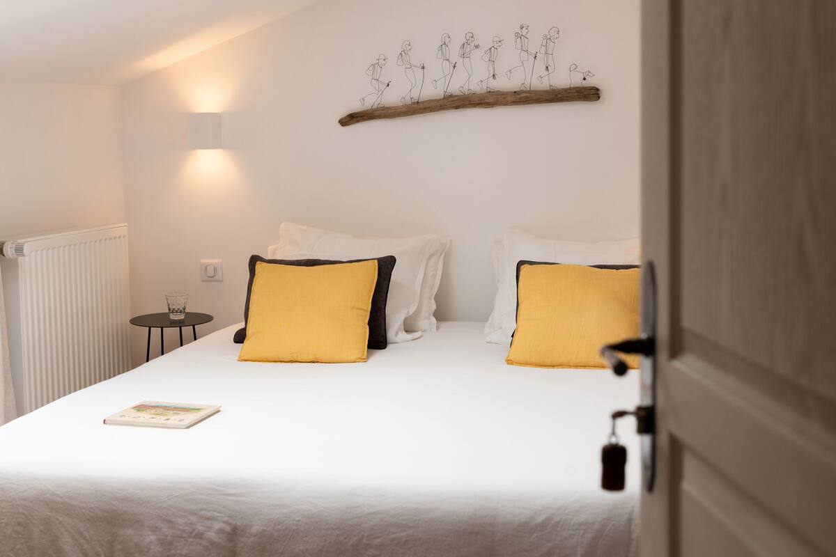 A bedroom with a bed, two pillows, a bedside table, a wall-mounted light, and a radiator.