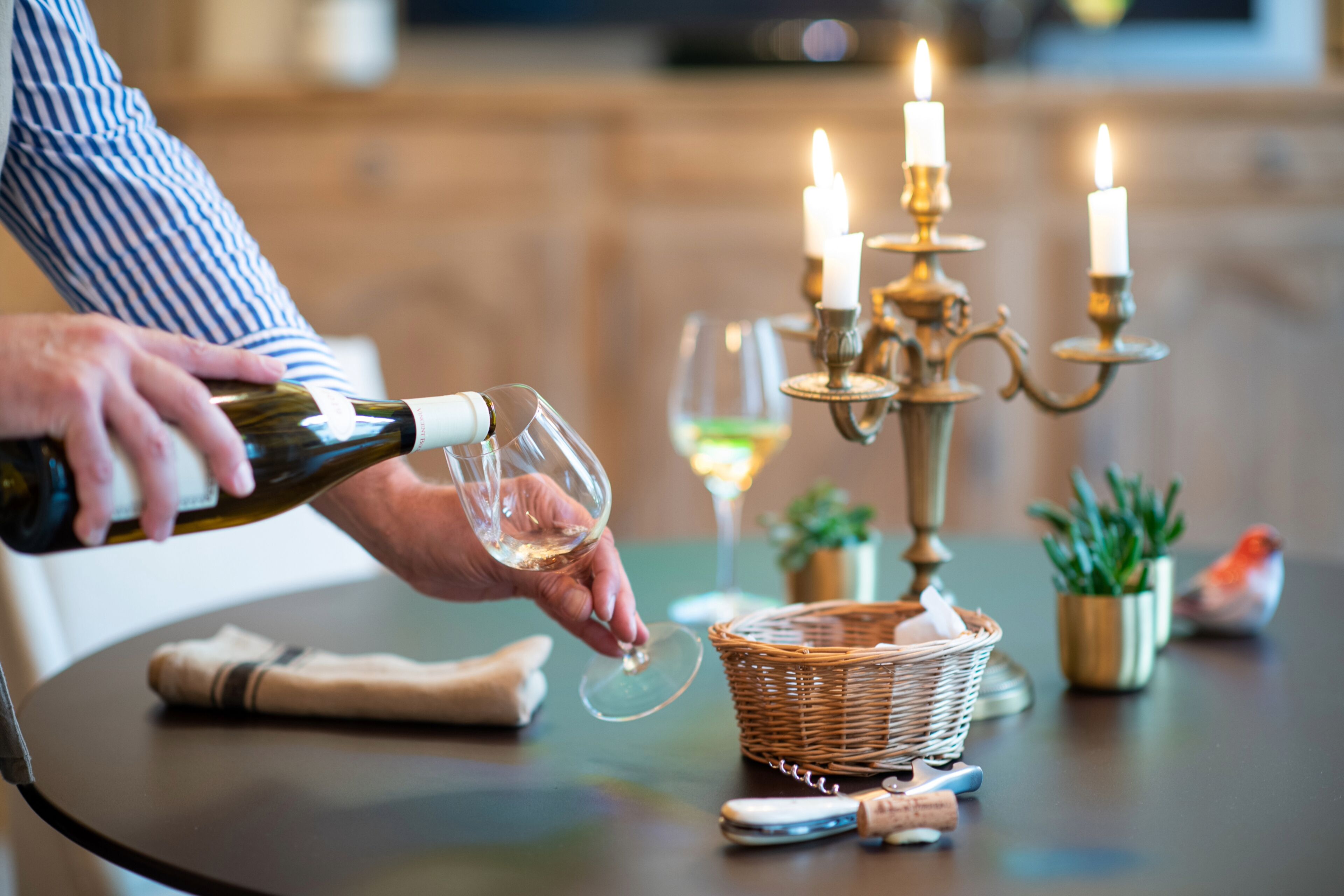 A person pouring wine into a glass, a candle holder with lit candles, and a basket on a table.
