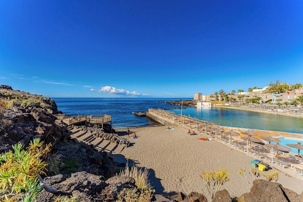 A beach with a rocky shoreline, a wooden walkway, and a pool area with umbrellas.