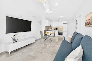 A modern living room with a large flat-screen TV, a sofa, a dining table with chairs, and a kitchen area in the background.