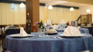 A dining table set with plates, cutlery, wine glasses, and a bottle of wine, with a vase of flowers and a decorative napkin on a blue tablecloth. - Hotel Cocoon (Pescasseroli)