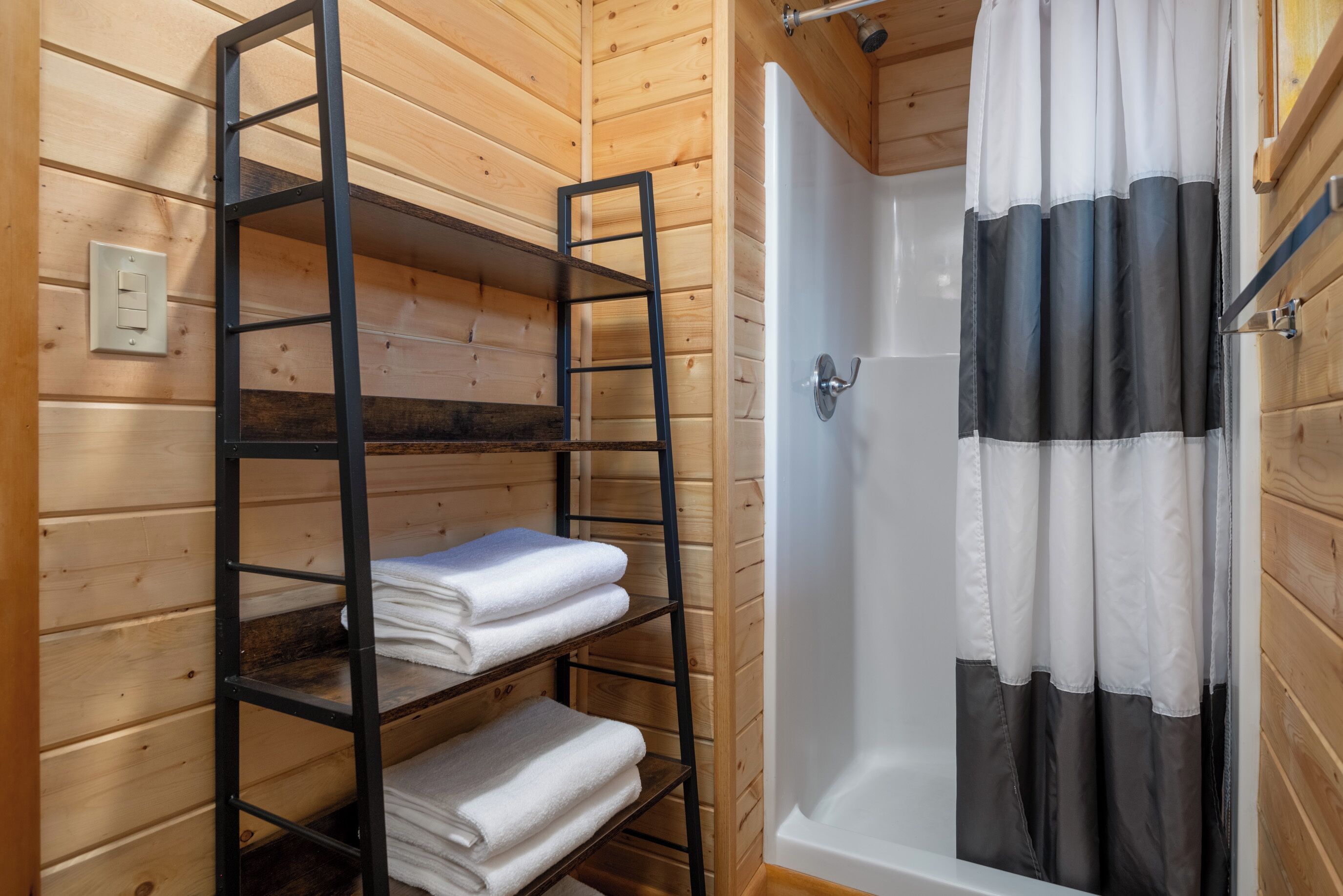 A wooden cabin bathroom with a wooden shelf holding towels, a shower with a black and white curtain, and a wooden door.