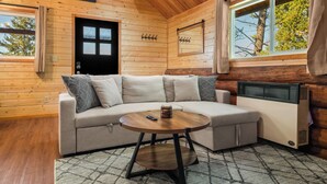 A cozy cabin interior with a wooden ceiling, a beige sofa, a round wooden table, and a large window with curtains. - Adventure Log Cabin with Expansive Views, Discovery Ski Hill nearby, walk to downtown. Perfect for couples and small families! (Philipsburg)