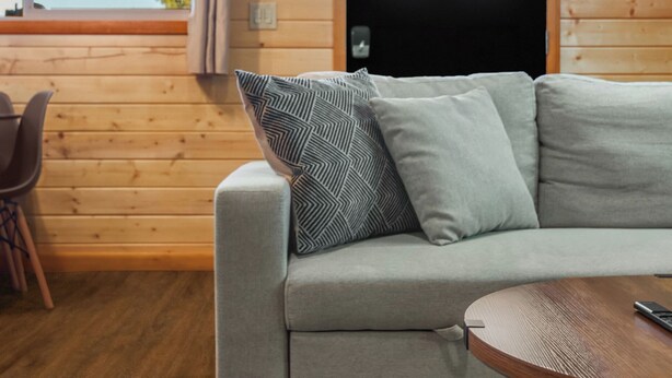 A cozy room with a wooden interior, a grey sofa with patterned cushions, a round wooden table, and a window with a view of the outdoors. - Adventure Log Cabin with Expansive Views, Discovery Ski Hill nearby, walk to downtown. Perfect for couples and small families! (Philipsburg)