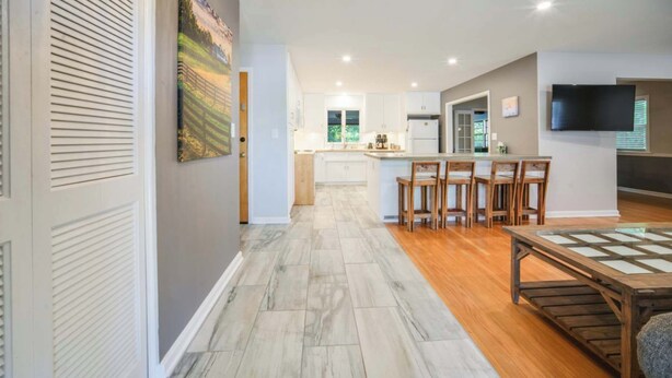 A modern kitchen with white cabinets, a wooden dining table, and a TV mounted on the wall.