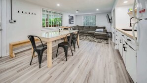 A modern kitchen with a dining table, chairs, and a sofa in a bright room with wooden flooring and large windows.