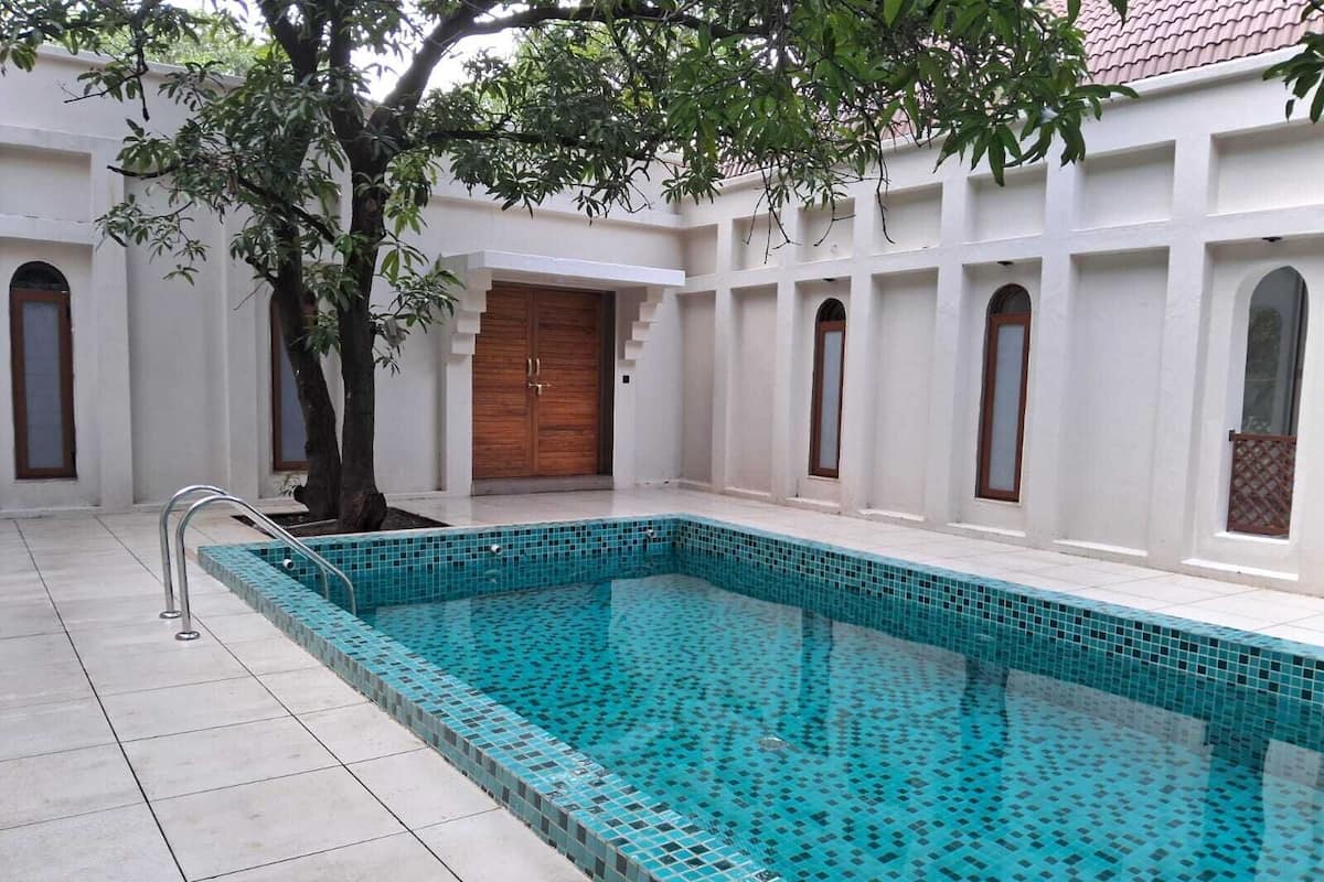 Uma piscina com borda azulejada, uma árvore e um edifício com janelas em arco e porta de madeira.