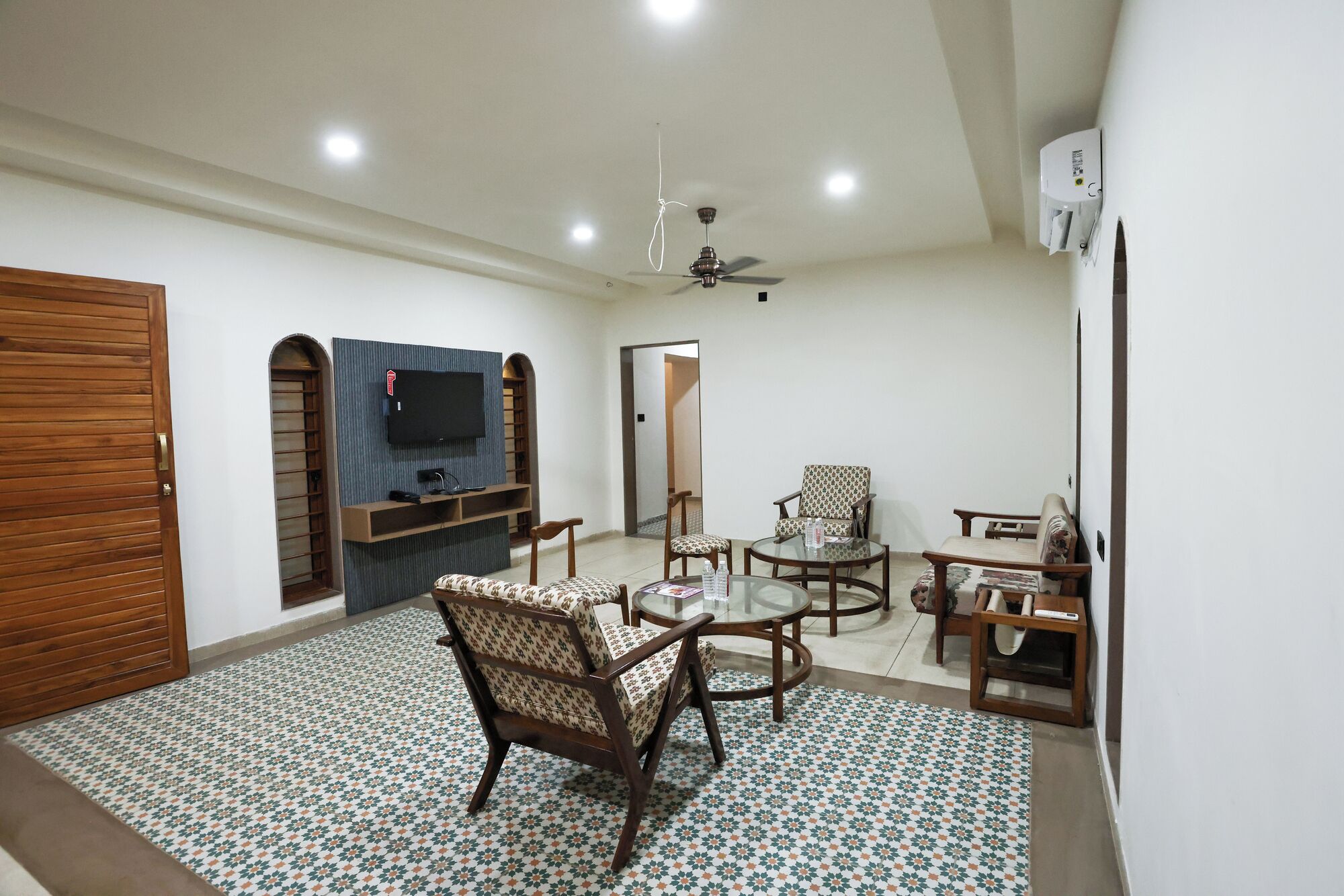 A living room with a TV mounted on a wall, a ceiling fan, a patterned floor, and a wooden door.