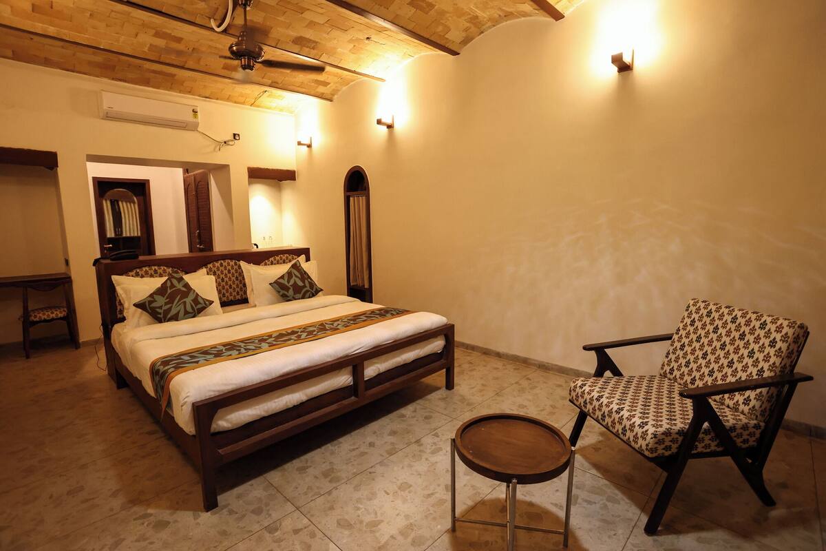 Quarto com cama, cadeira, mesa pequena e ventoinha no teto.