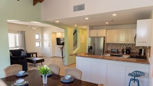 A modern kitchen with white cabinets, a stainless steel refrigerator, and a built-in oven. There is a dining area with a dark wooden table set for four, surrounded by wicker chairs. - Palm Paradise at Tierra Del Sol - 2 bedroom/2 full baths and 2 1/2 baths is just waiting for you to enjoy some sun and fun! (Noord)