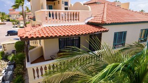 A two-story building with a red-tiled roof, a balcony with white balustrades, and a view of a street with parked cars. - Palm Paradise at Tierra Del Sol - 2 bedroom/2 full baths and 2 1/2 baths is just waiting for you to enjoy some sun and fun! (Noord)
