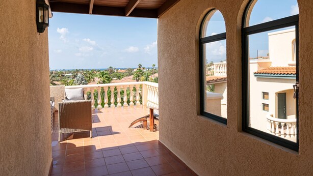 A balcony with a wicker chair and a table, overlooking a coastal view with buildings and palm trees. - Palm Paradise at Tierra Del Sol - 2 bedroom/2 full baths and 2 1/2 baths is just waiting for you to enjoy some sun and fun! (Noord)