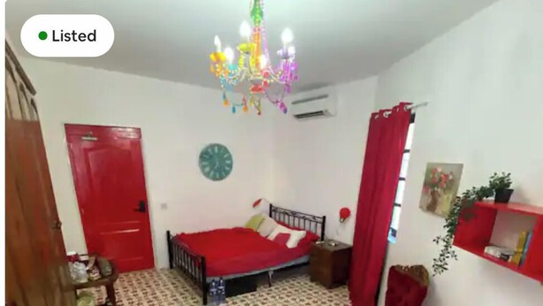 A bedroom with a bed, a green rug, a red door, a clock, a chandelier, and a small shelf with items. - Maleth House (Naxxar)