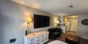 A modern living room with a flat-screen TV, a sofa, a coffee table, and a kitchenette in the background.