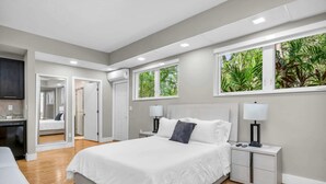 A modern bedroom with a large bed, two bedside tables, and a nightstand. There are two windows with a view of greenery outside.