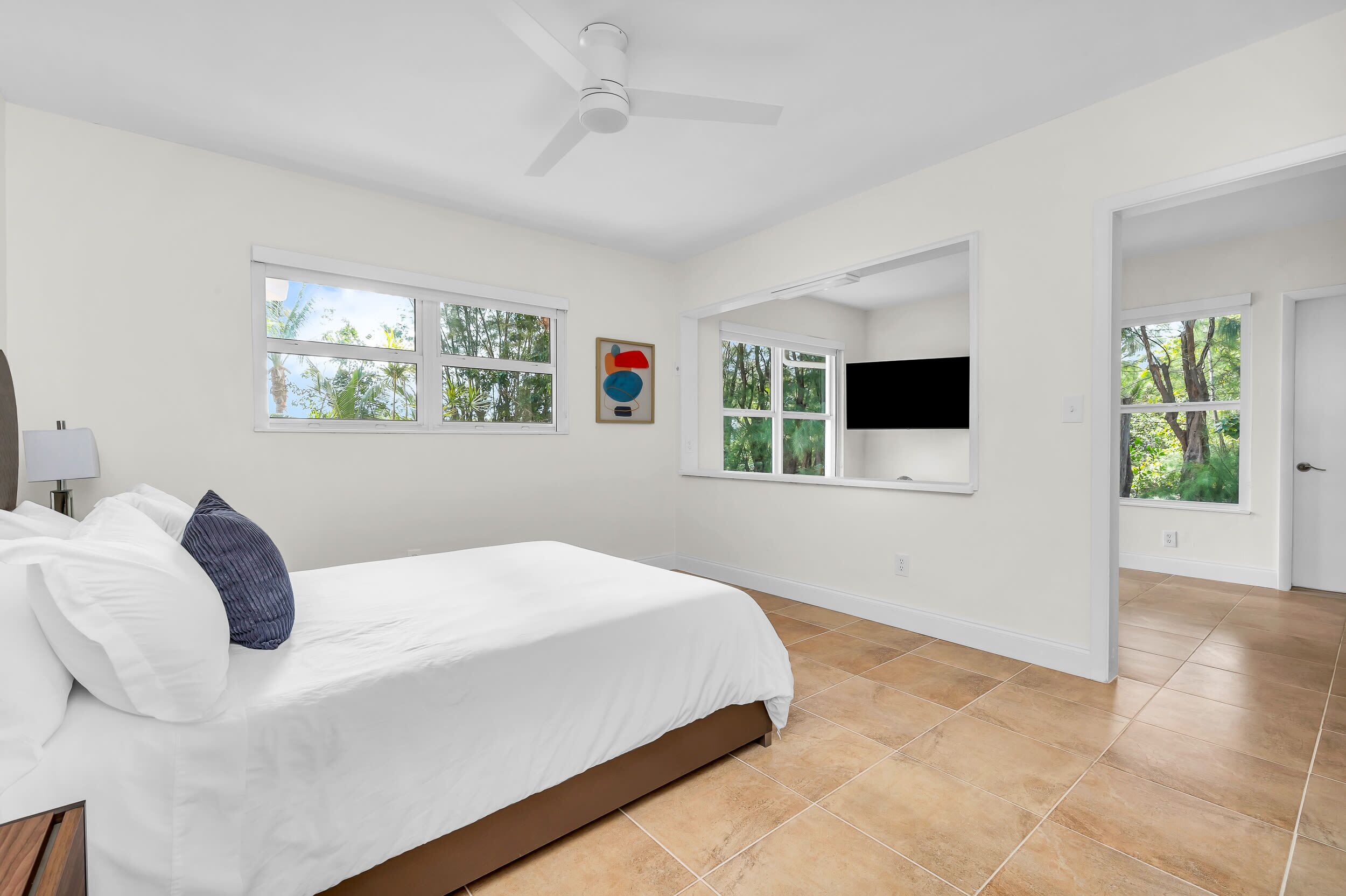 Un dormitorio con cama, paredes blancas, ventilador de techo, televisor montado en la pared y ventanas con vista a árboles.