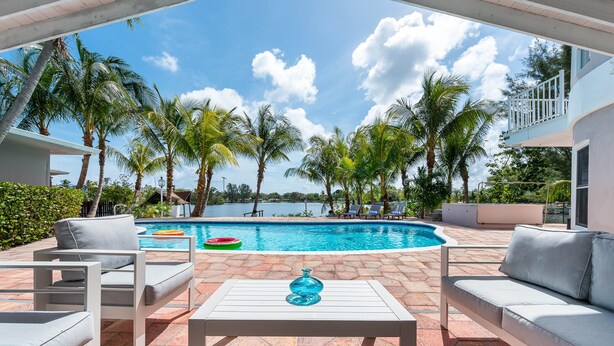 A poolside area with white lounge chairs, a white table, and a view of the pool and palm trees.