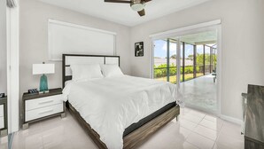 A bedroom with a large bed, a nightstand with a lamp, a ceiling fan, and a view of the outdoors through a sliding glass door.