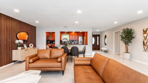 A modern living room with a brown leather sofa, a dining area with a wooden table and chairs, and a kitchen in the background.