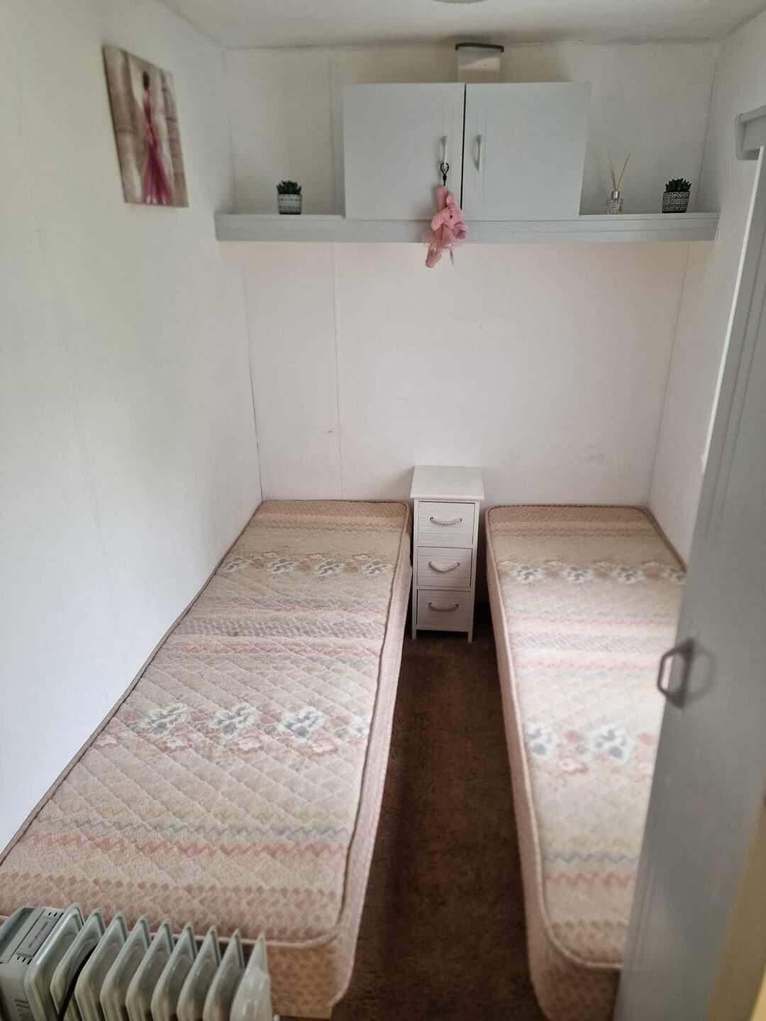Una habitación pequeña y funcional con dos camas individuales, un armario blanco decorado con motivos rosas y un radiador al pie de las camas.