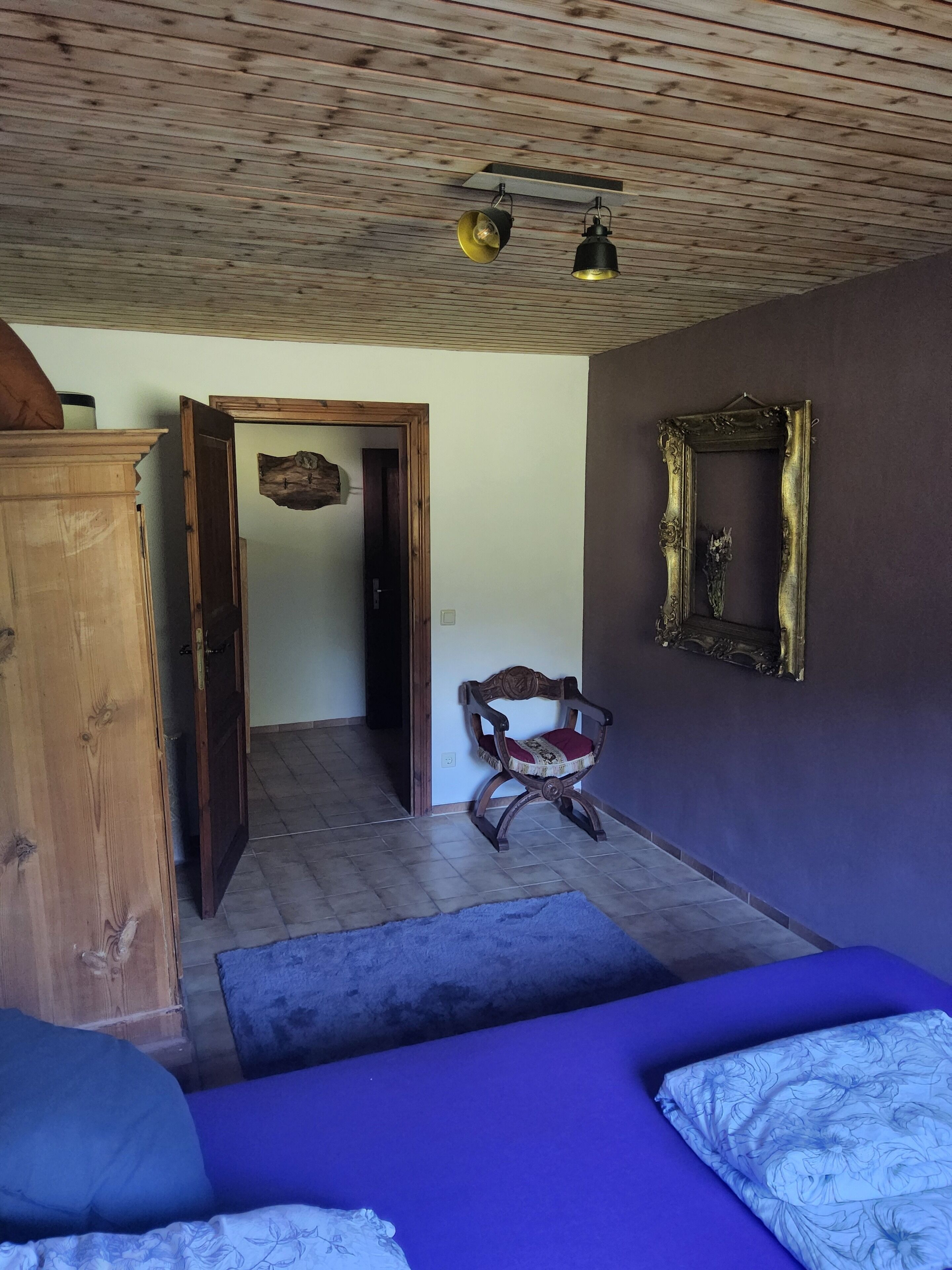 A room with a wooden ceiling, a bed with blue bedding, a chair with a red cushion, a wooden cabinet, and a mirror on the wall.