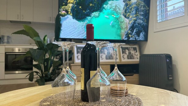 A kitchen with a TV displaying a scenic landscape, a bottle of wine, and three wine glasses on a woven placemat. - SYDNEYS LEADING NO.1 STAY (Coogee)