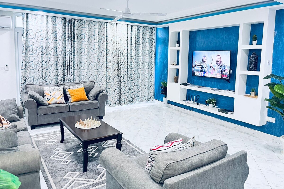 A modern living room with a grey sofa set, a coffee table, a TV mounted on a blue accent wall, and patterned curtains.