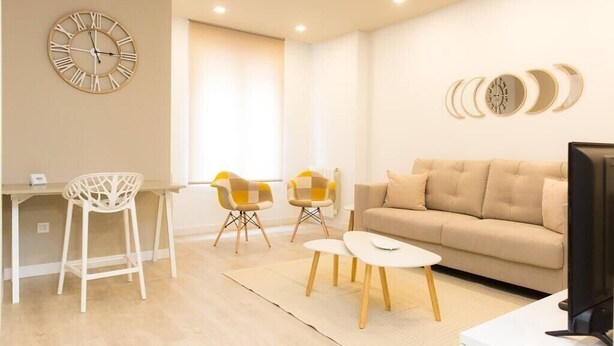 A modern living room with a beige sofa, a white coffee table, and two chairs. A large wall clock and a series of moon phase wall art are visible.