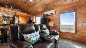A cozy cabin interior with a leather sofa, wooden walls, a dining area, and a large window offering a view. - Valley View Cottage (Omaha)