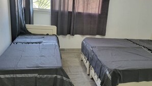 Ein kleines Zimmer mit zwei Einzelbetten, einem Fenster mit Vorhängen und einer schlichten weißen Wand.