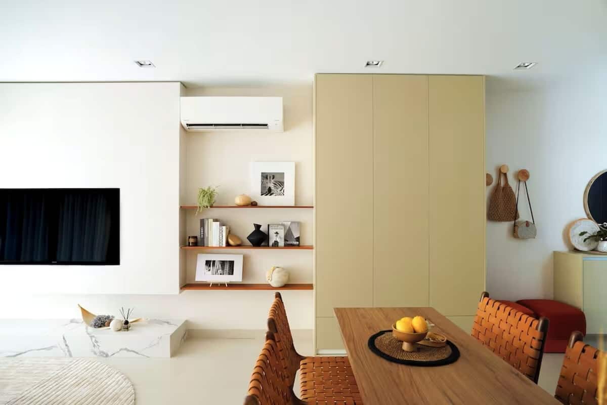 A modern living room with a wooden dining table, a sofa, and a TV mounted on the wall. There are shelves with books and decorative items, and a large cabinet.