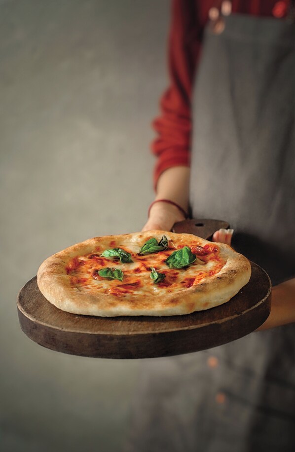 A pizza on a wooden board held by a person wearing a red shirt and gray apron.