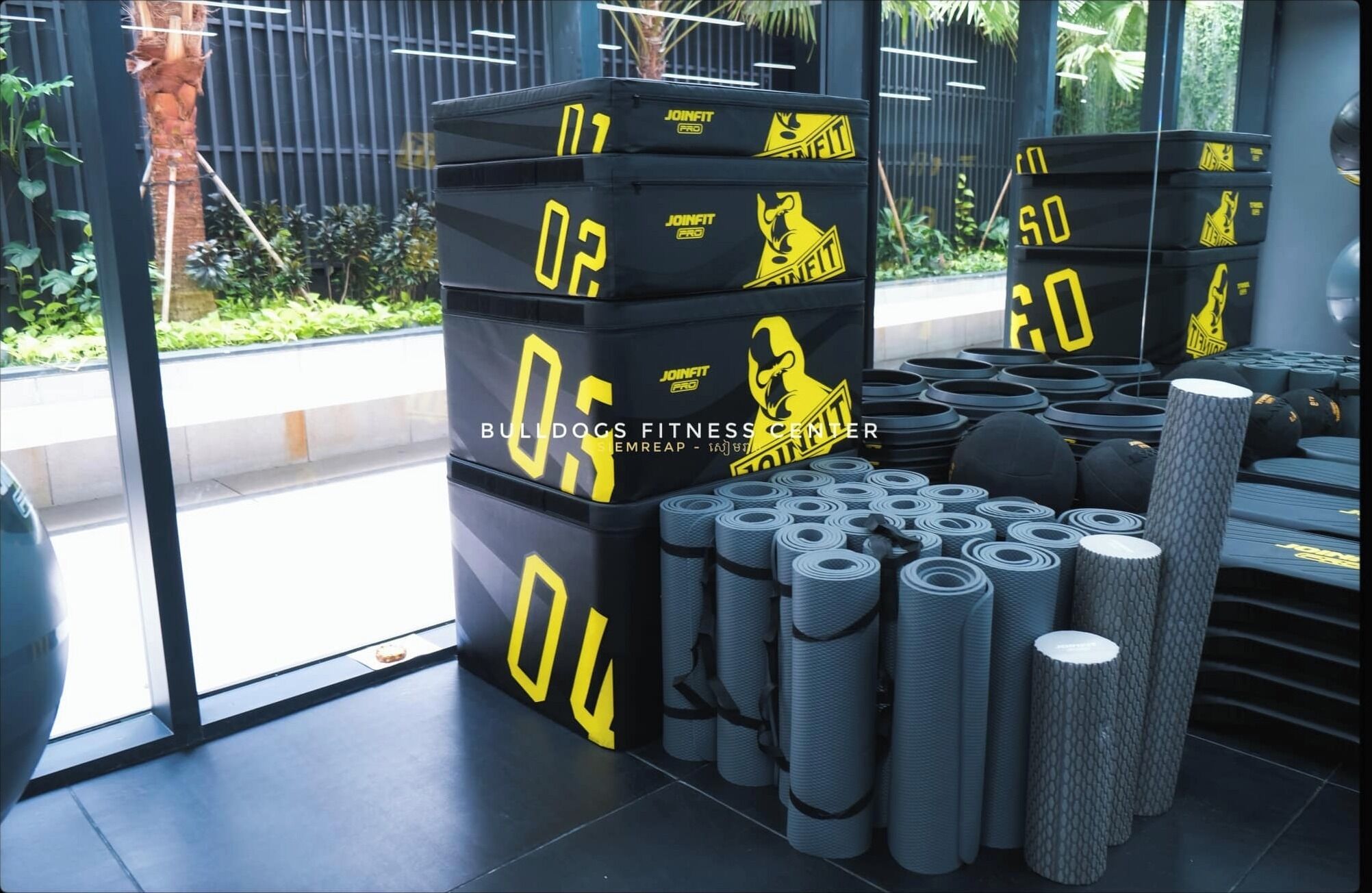 A fitness center with stacked mats labeled with numbers and a logo, and multiple rolled-up mats on the floor.