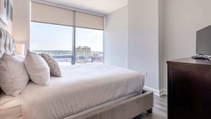 Een moderne hotelkamer met een groot bed, een donkere houten dressoir en stadszicht door ramen van vloer tot plafond.