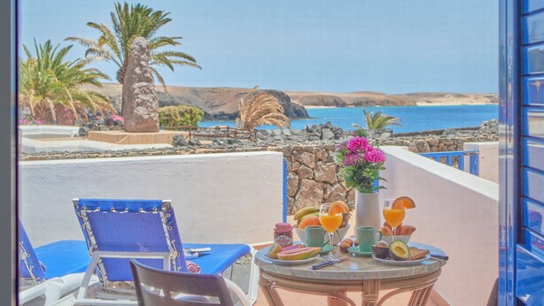 Un balcón con una mesa dispuesta para el desayuno, con vista a un paisaje costero con palmeras y un mar azul claro.