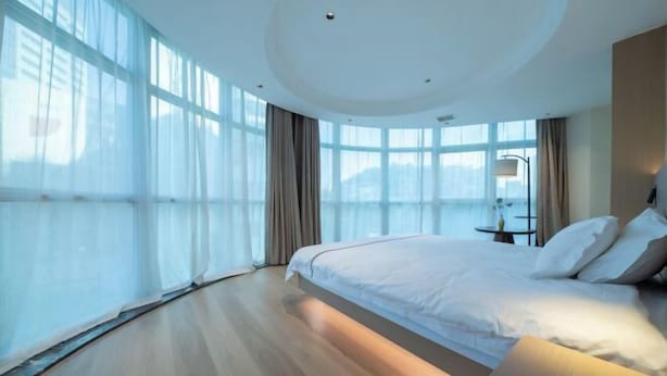 A modern hotel room with a large bed, a bedside table, and a view of the city through large windows. - MONI HOTEL ARTS (Jingdezhen)