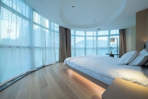 A modern hotel room with a large bed, a bedside table, and a view of the city through large windows.