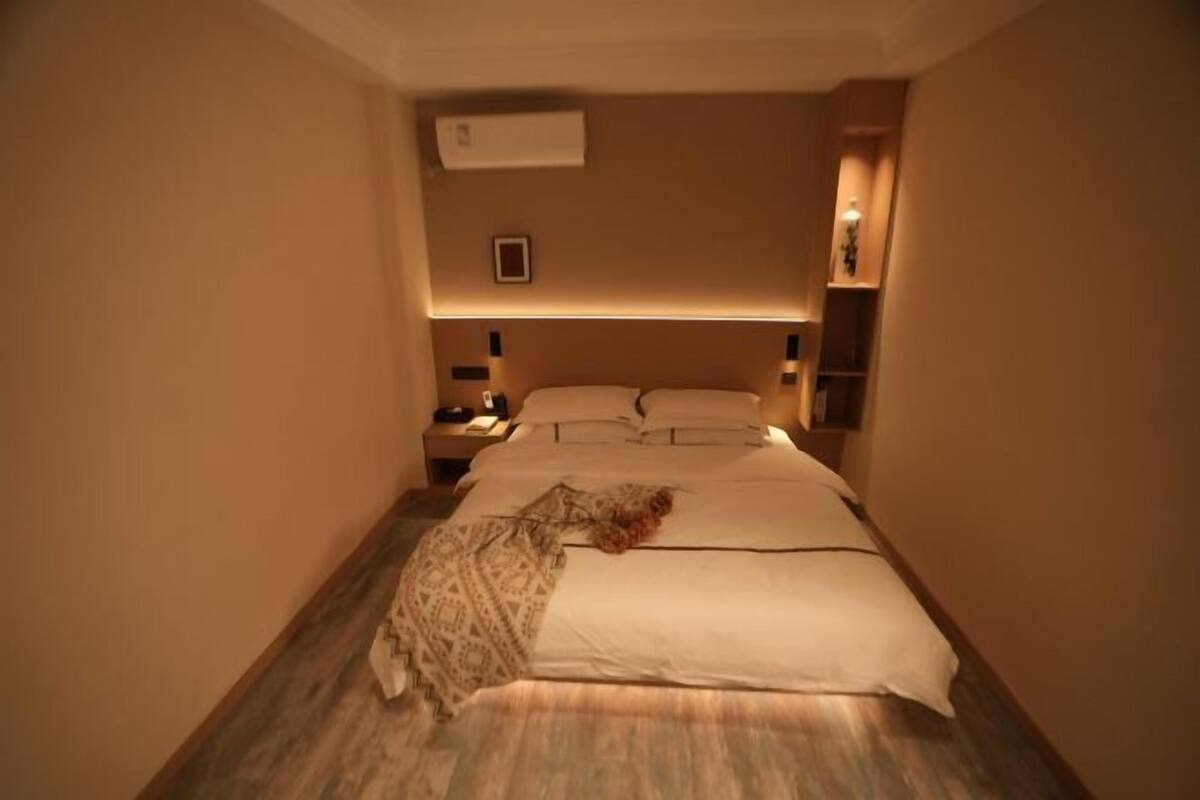A hotel room with a bed, bedside tables, a nightstand, a wall-mounted air conditioner, and a small shelf with a lamp.