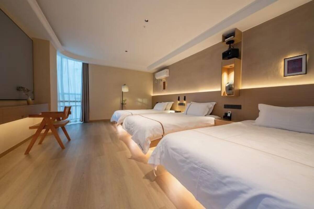 A hotel room with a wooden floor, three beds, a desk with chairs, and a large window.