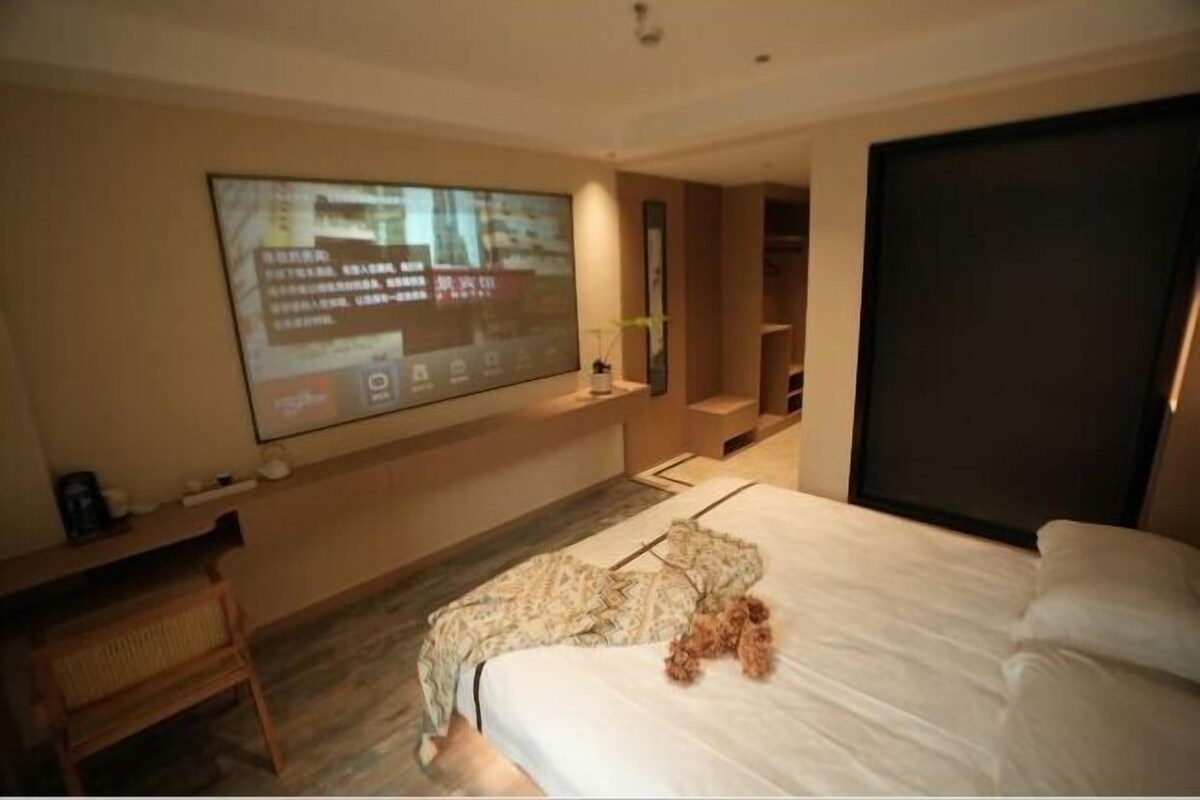 Habitación de hotel moderna con una cama grande, un televisor de pantalla plana montado en la pared, una silla de madera y una pequeña mesa con una planta.