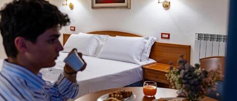 A hotel room with a bed, bedside lamps, a painting, a table with breakfast, and a vase of flowers.