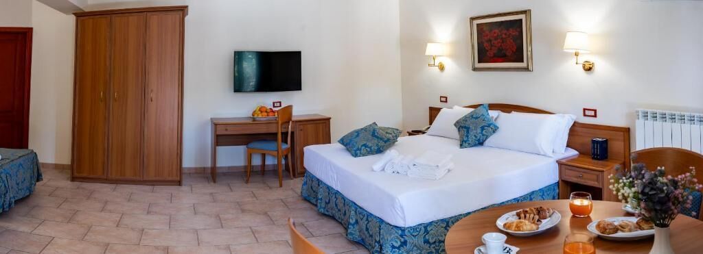 A hotel room with a bed, a desk, a chair, a television, and a dining table with breakfast items.