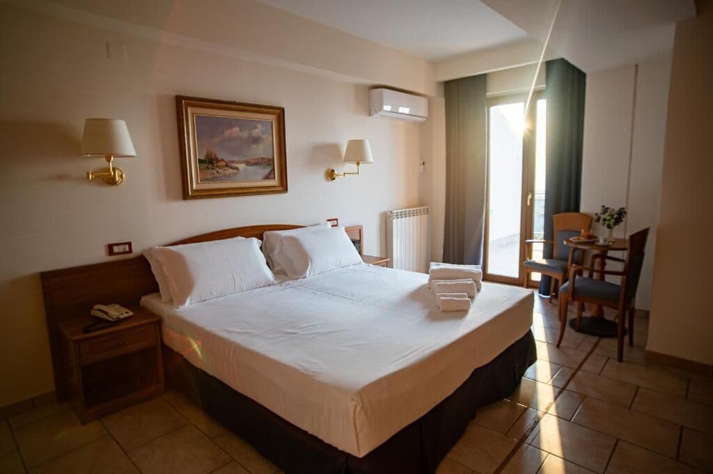 Habitación de hotel con una cama grande, lámparas de noche, un cuadro, un escritorio con una silla y vistas al exterior.