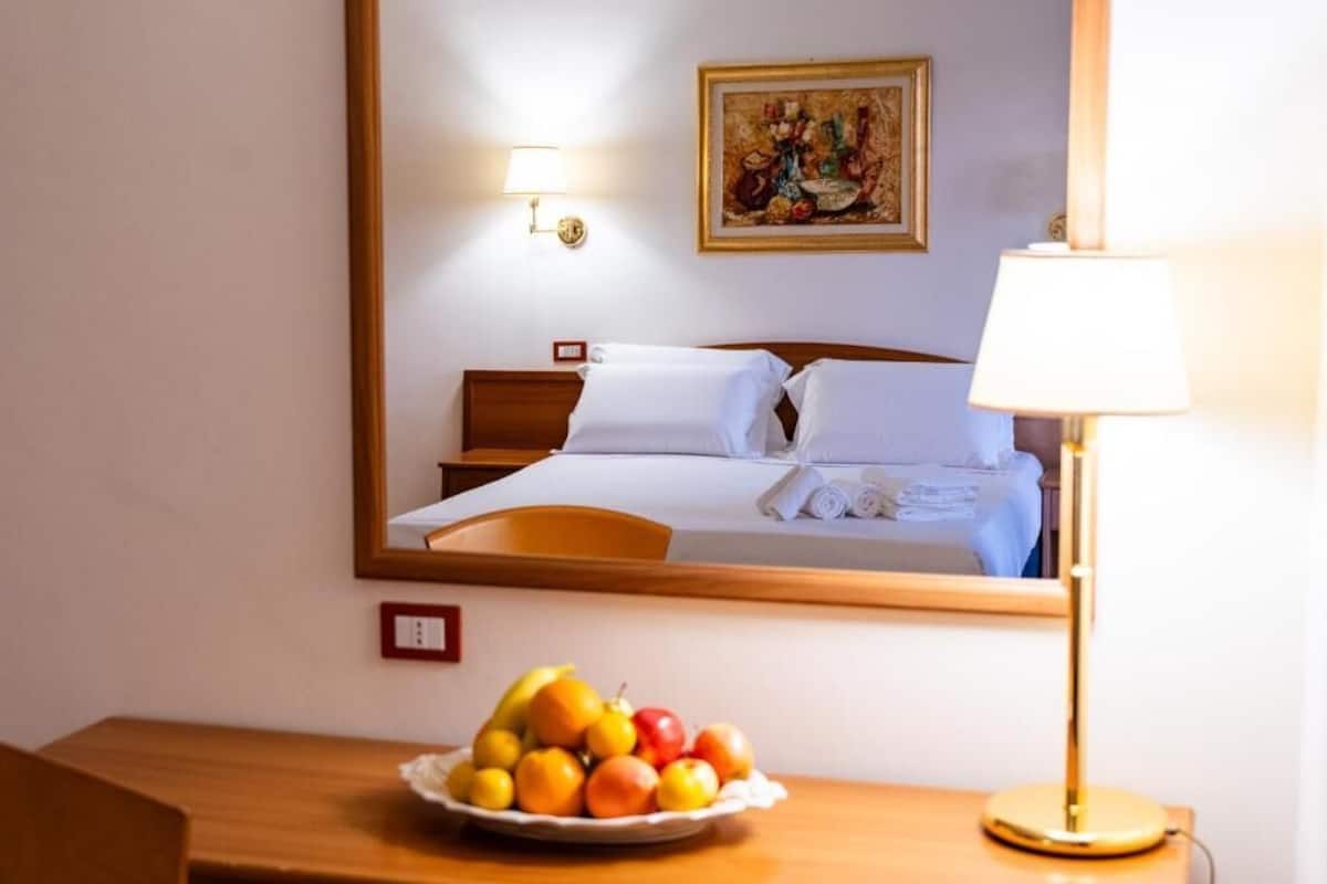 Una habitación de hotel con una cama, una mesita de noche con un frutero, una lámpara y un cuadro en la pared.