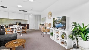 A modern living room with a sofa, coffee table, TV, and dining area with chairs and a table. - Coastal Lights I Pool, Gym & Parking@HarbourLights (Cairns City)