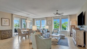 A spacious living room with a dining area, large windows, a ceiling fan, and a television.