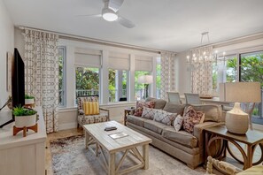 A spacious living room with a sofa, coffee table, and a TV. There are large windows with curtains, a ceiling fan, and a chandelier. - Bayview Luxury: Sandestin Retreat, Resort Perks (Miramar Beach)