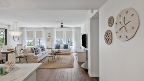 Ein modernes Wohnzimmer mit einer Couch, einem Couchtisch und einem Fernseher. Große Fenster und ein Deckenventilator sind vorhanden.